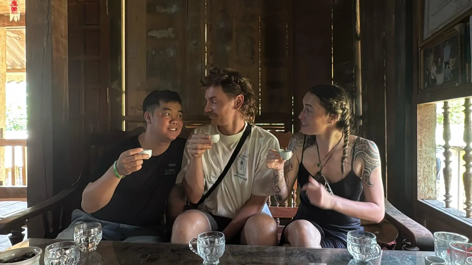 A guide and two guests raising small cups of rice wine inside a wooden house